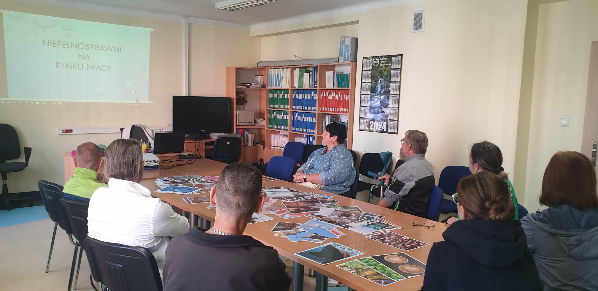 Fotografia wykonana podczas warsztatów realizowanych 18.09.2024 r. Przedstawia salę spotkań grupowych a w niej kilka osób siedzących przy wspólnym stole. 