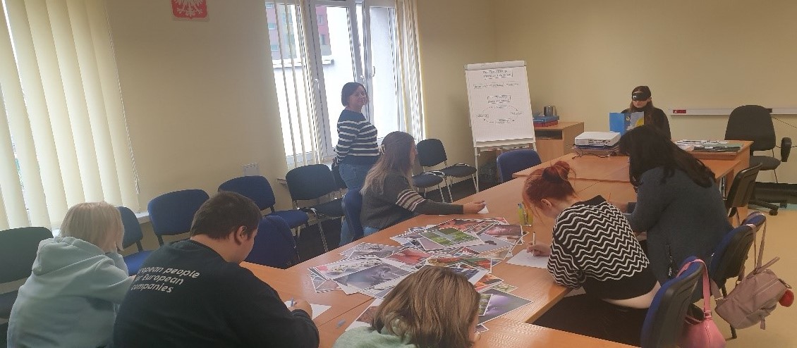 Fotografia wykonana podczas warsztatów realizowanych 07.11.2024 r. Przedstawia salę spotkań grupowych a w niej kilka osób siedzących przy wspólnym stole. 